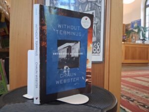 Image of the cover of the book Without Terminus: untraining an archive by chaun webster is the foreground. The book is on a stand, positioned on a library step-stool. In the background there is a poster by Ricardo Levins Morales that invokes the 1968 Civil Rights campaign phrase in Memphis, Tennessee, with a crowd of Black men holding signs that read, “I am a man”. The background includes a library bookshelf, part of the central desk at the East Side Freedom Library, and a tall window typical of the Carnegie style architecture of the library.